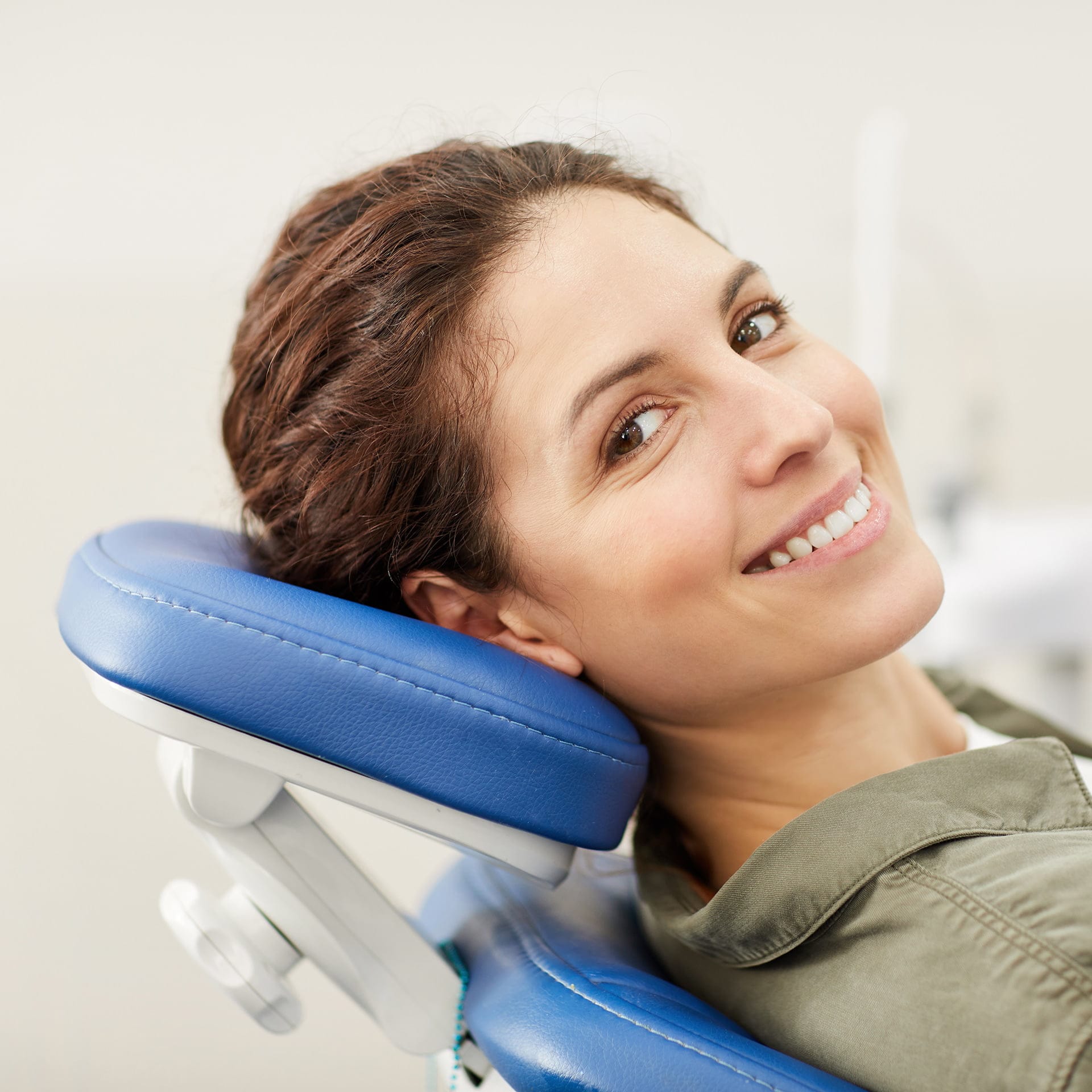 A smiling woman with brown hair reclining comfortably in a dentist's chair, exhibiting a relaxed and positive demeanor. She's wearing a casual olive green jacket and the chair is upholstered in blue, indicating a clean and professional dental care environment.