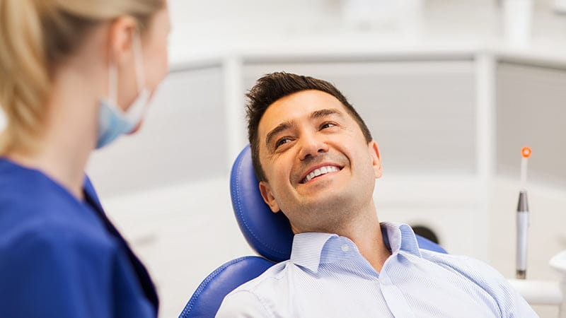 A male patient in a dental chair smiles up at a dental professional, conveying trust and comfort. The patient is dressed in a crisp blue shirt, and the dental professional, partially visible and wearing a blue uniform and face mask, appears engaged and attentive. The modern dental office background features clean lines and dental equipment, highlighting a professional atmosphere.