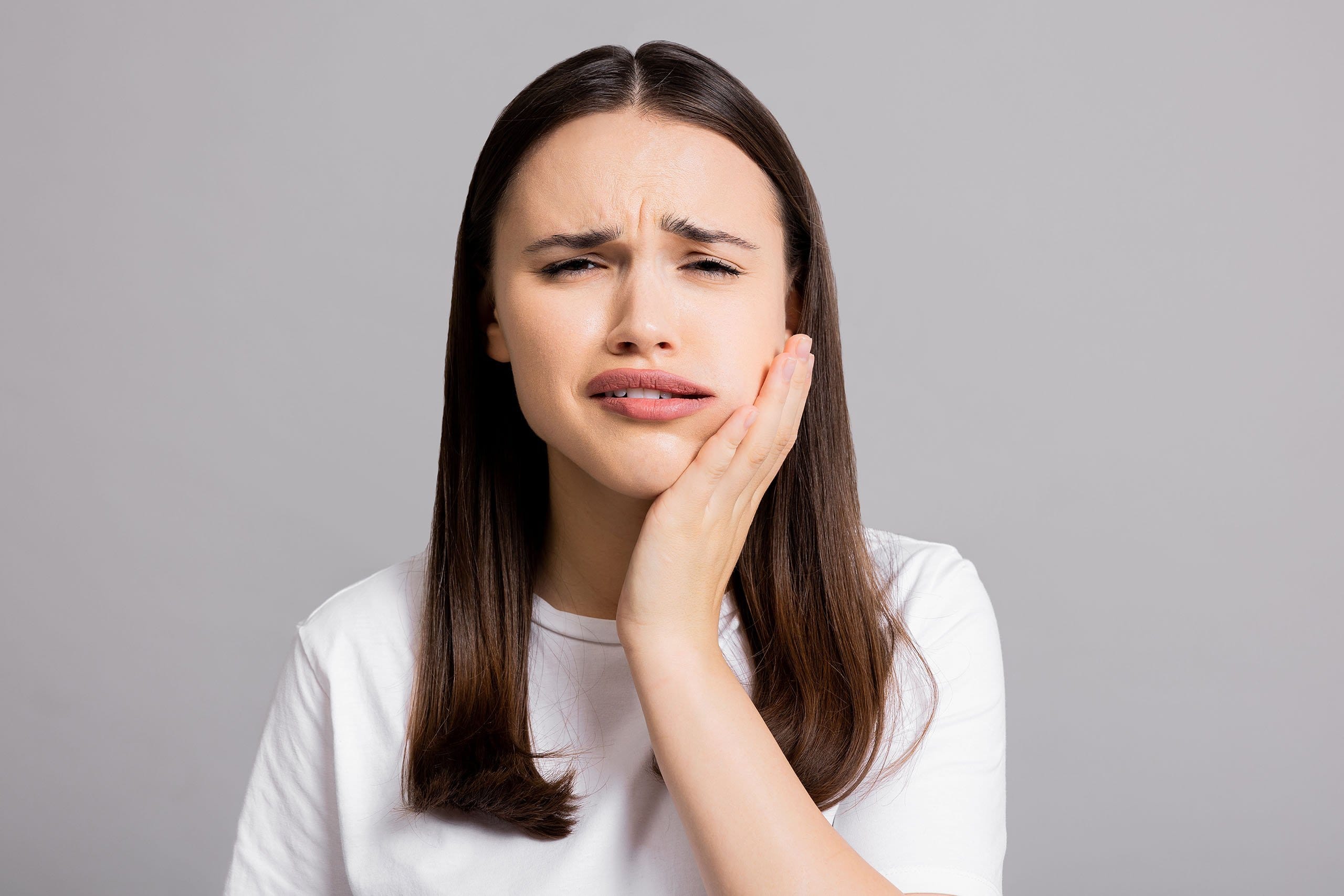 A woman in a white shirt with a pained expression, pressing her cheek and closing her eyes slightly as if experiencing tooth pain from a dental abscess, against a neutral gray background.