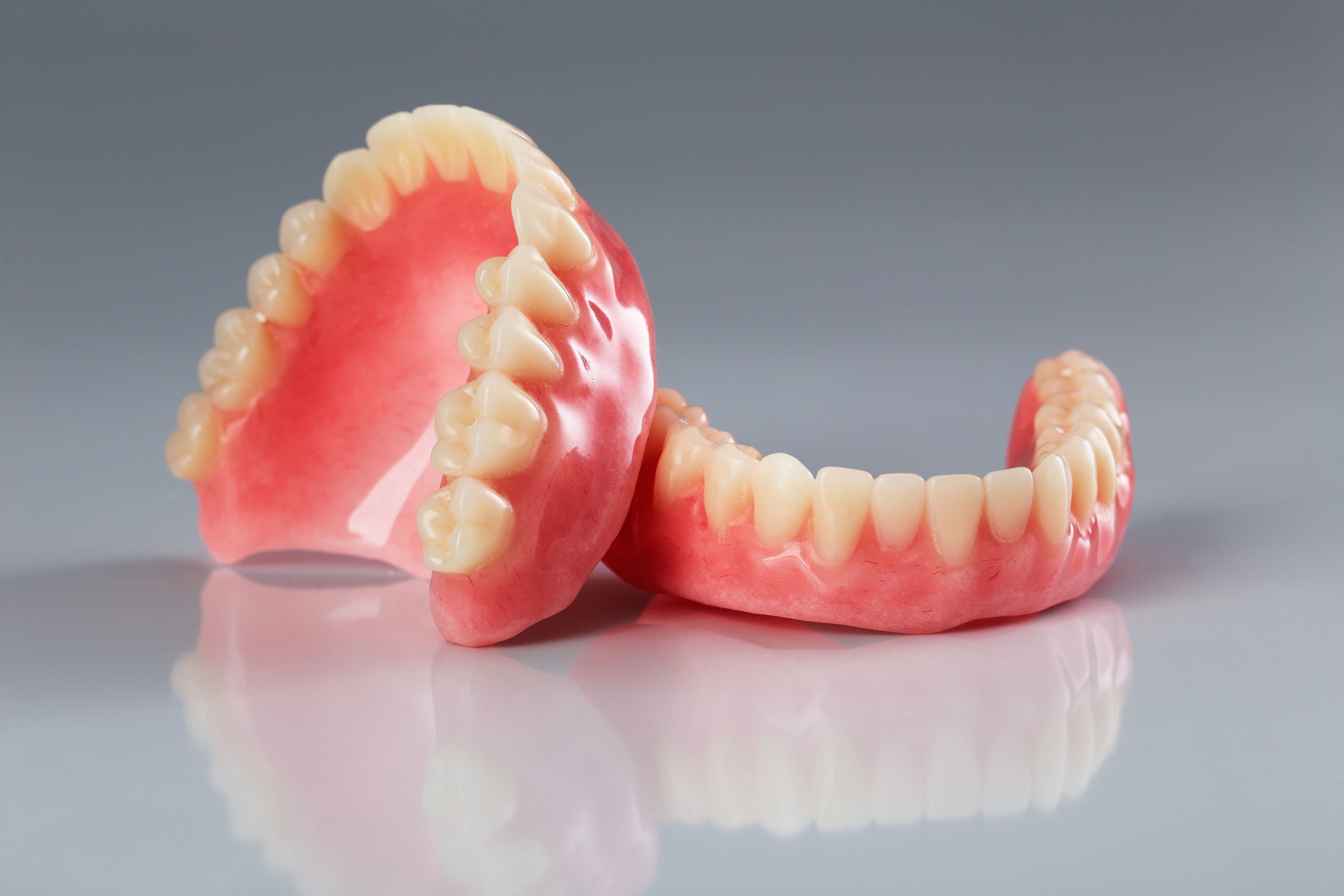 A pair of upper and lower full dentures with pink gums and white teeth, displayed side by side on a reflective surface with a soft gray background.