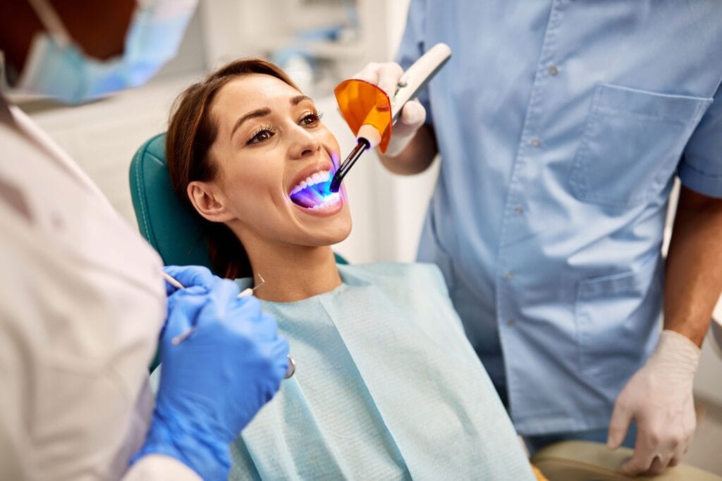 A smiling woman in a dental chair receiving a a dental filling treatment with a blue curing light, as a dentist in a blue uniform holds a dental instrument and an orange protective shield.