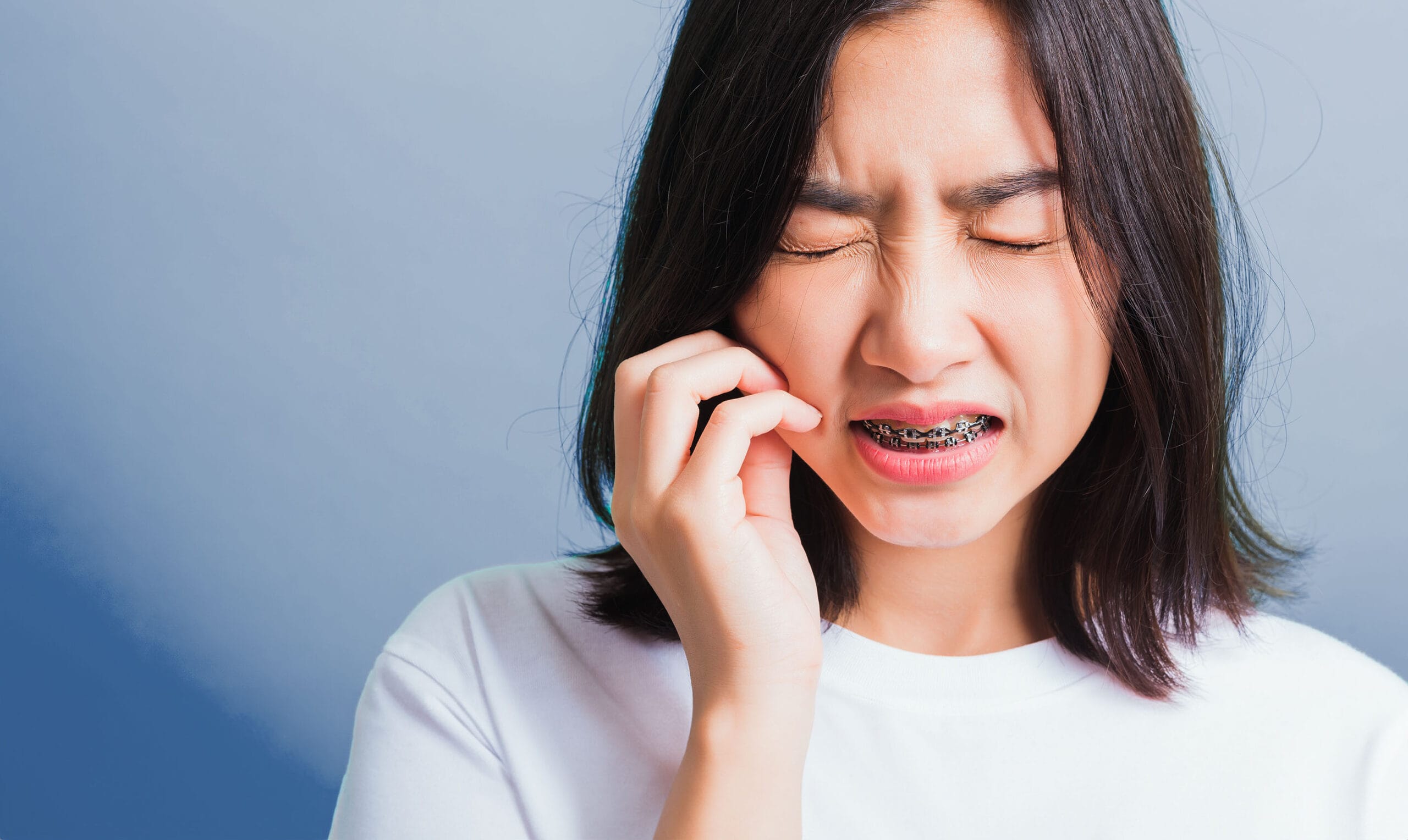 A young woman with braces on her teeth frowning and touching her cheek, appearing to be in discomfort due to an orthodontic injury, set against a soft blue background.