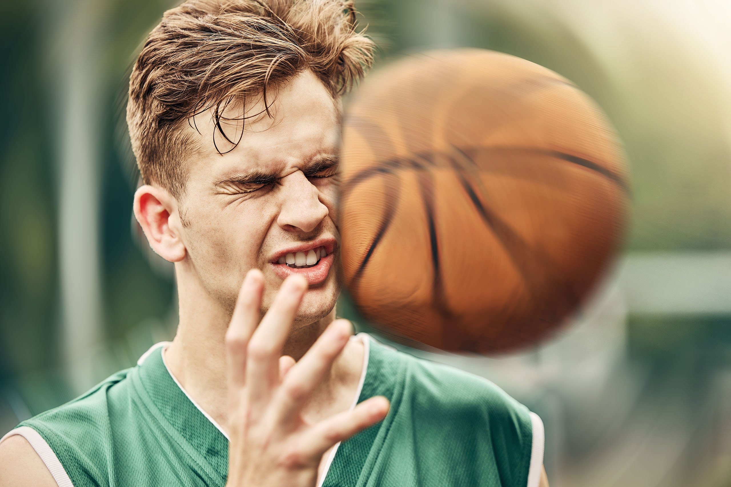 A man in a green jersey wincing and recoiling as a basketball approaches his face, capturing a moment of impact and potential sports injury during an outdoor game.