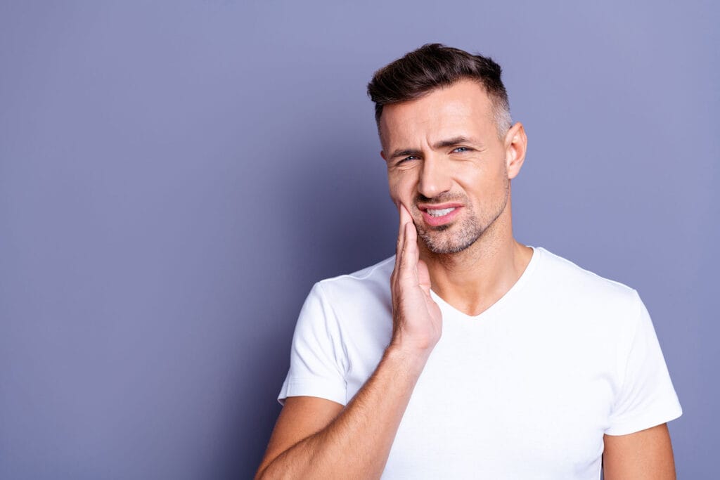 Man holding his cheek with a pained expression, indicating discomfort on one side of his jaw.