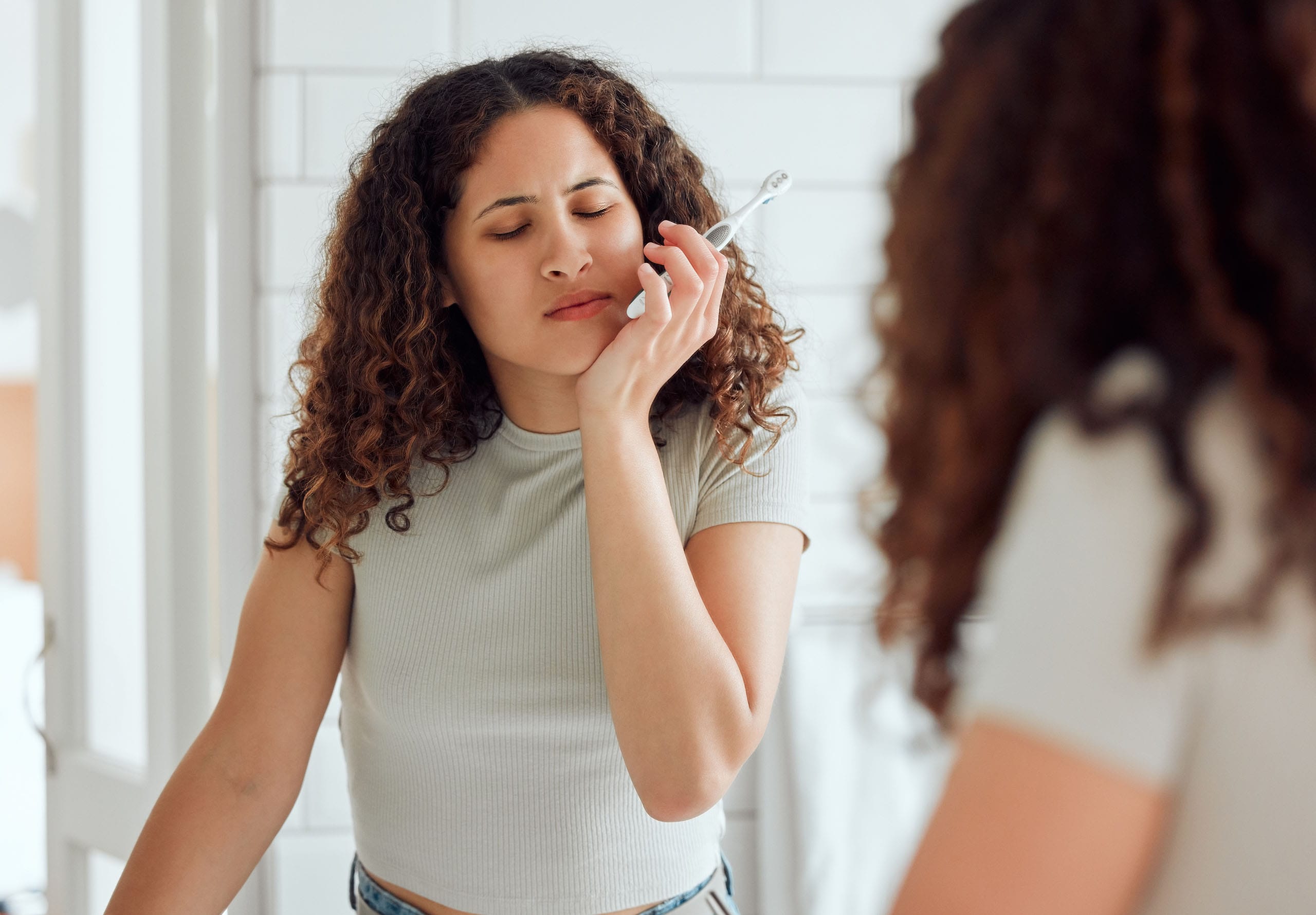 Woman touching her cheek while holding a toothbrush, appearing to experience tooth pain in a bathroom.