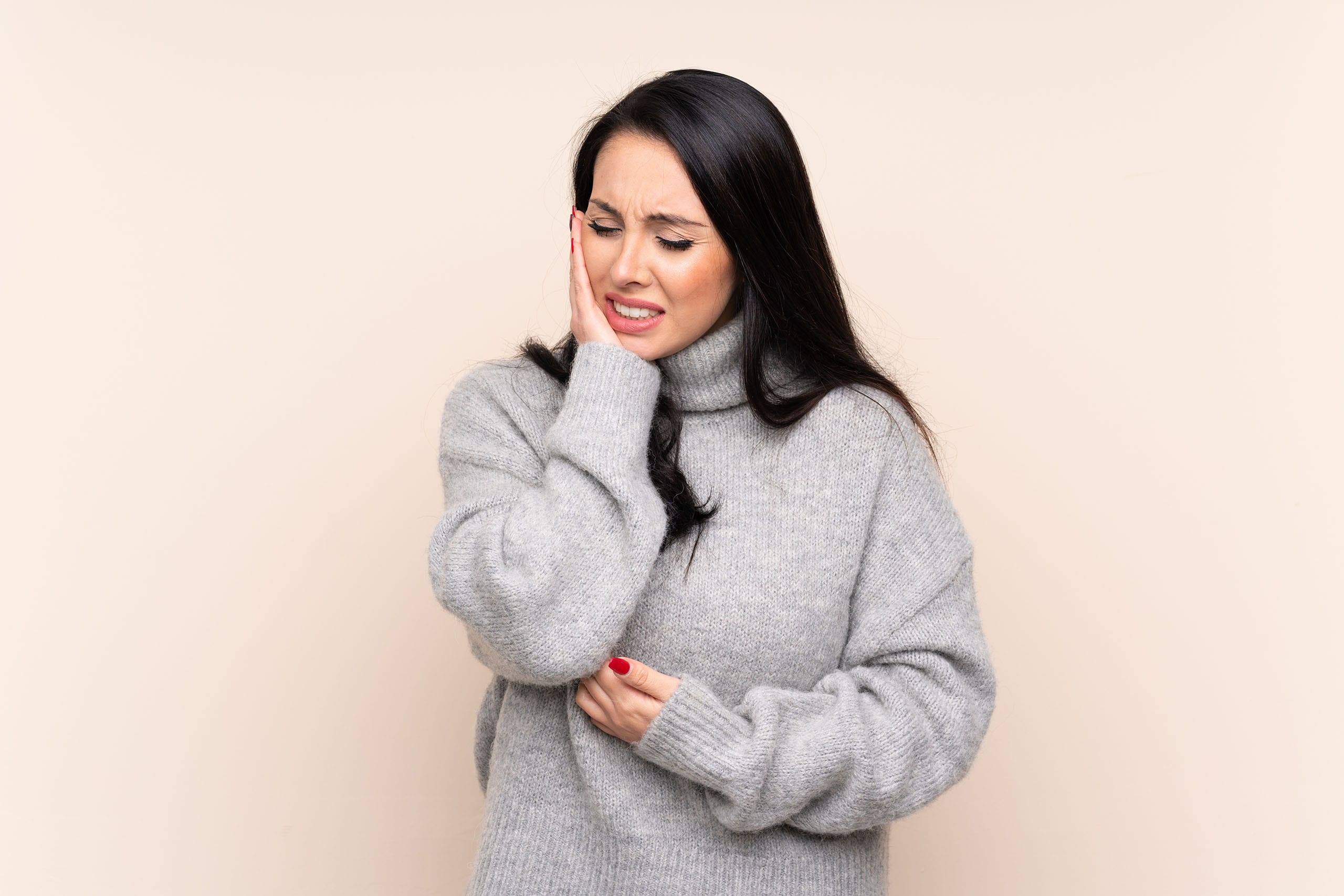 Woman in a gray sweater holding her jaw with a pained expression against a neutral background.