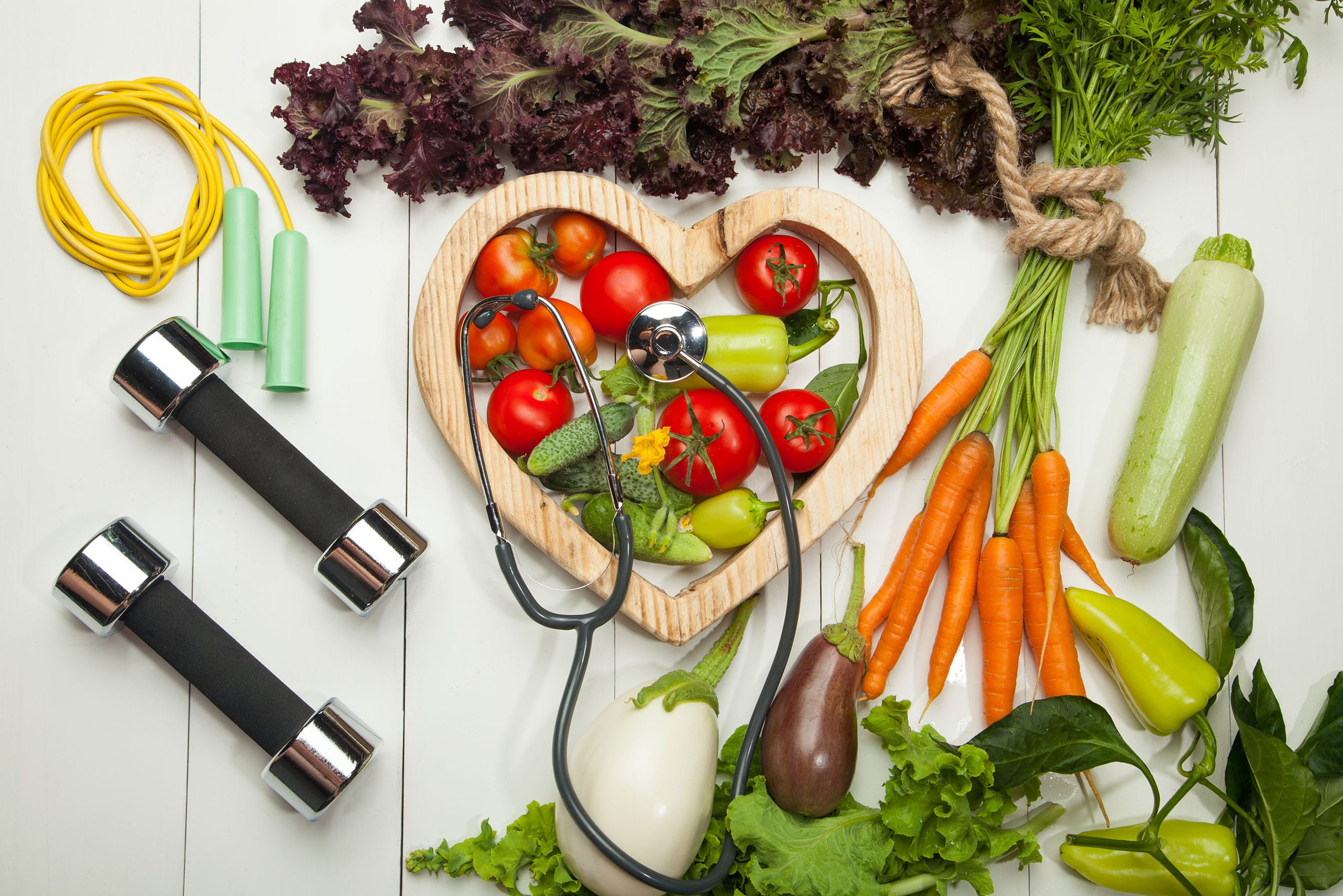Fresh vegetables in a heart-shaped wooden bowl with stethoscope, dumbbells, and jump rope on white table.