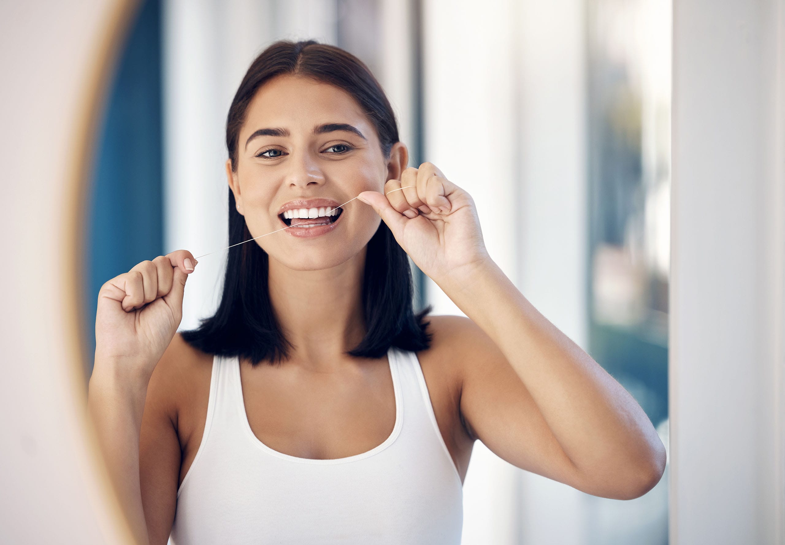 Woman flossing teeth in bathroom mirror, demonstrating daily oral hygiene for healthy gums