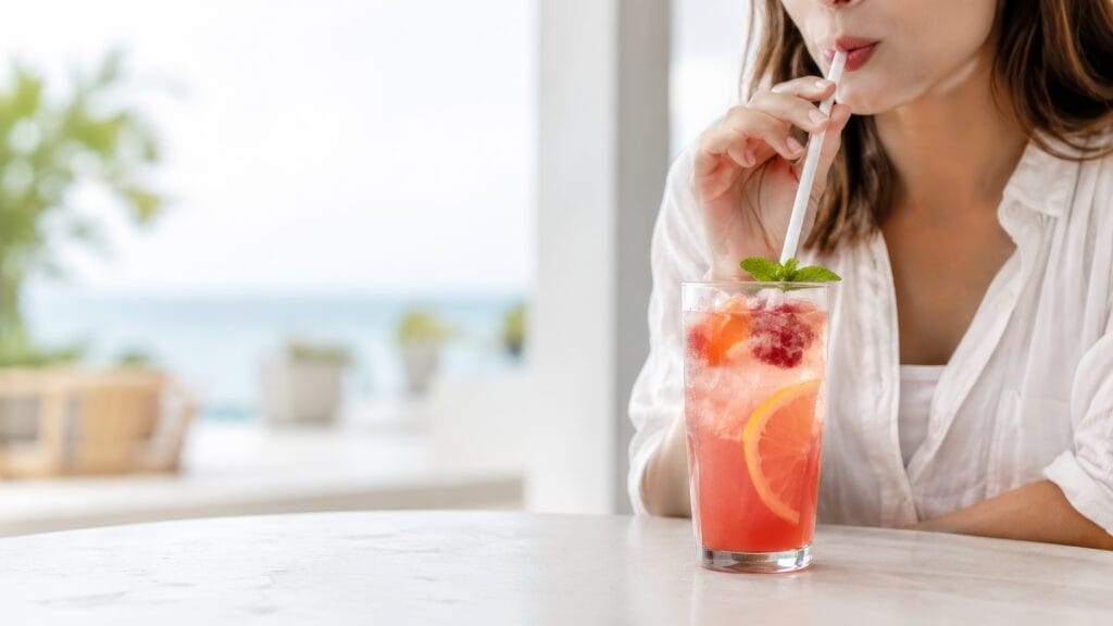 Adult sipping a pink citrus drink through a straw at a bright table, highlighting a tooth-friendly way to enjoy sugary or acidic beverages.
